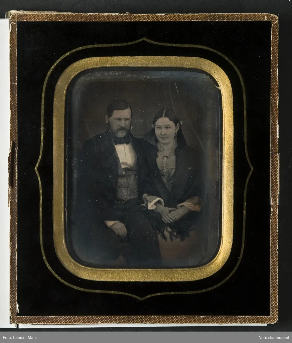 Porträtt av Adolf Drakenberg (1827–1896) och hans hustru Augusta Adler (1834–1866), knäbild, sittande. Han, till vänster, med mustasch och polisonger, klädd i svart frack eller bonjour. Hon, till höger, med mittbenat hår, ljus klänning med vita manschetter och svart sjal. Dagerrotyp / daguerreotyp helt glasad. Passepartout i svart färg målad direkt på glaset. Svart bakstycke av papp med två påklistrade vita etiketter med handkriven text: "Johan Adolf Drakenberg/f. 1827 +1896/hans hustru Augusta Adler f. 1834 +1866" och "Patron/Adolf Drakenberg och /hans hustru Augusta Adler/porträtt taget 1852". Nordiska museet inv.nr 143947. 
-
Portrait of Adolf Drakenberg (1827–1896) and his wife Augusta Adler (1834–1866). Third-plate daguerreotype dated 1852.