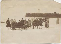 Ti personer ved hest og slede foran et hus eller hytte