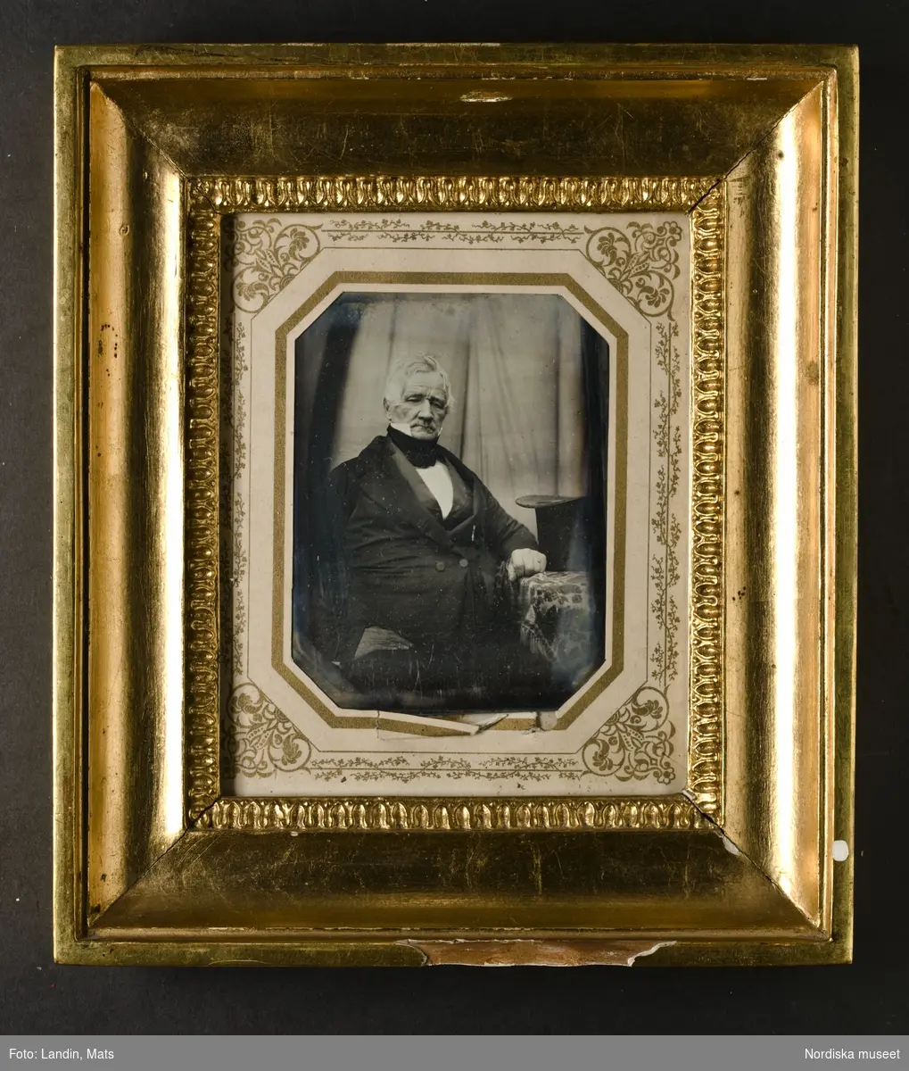 Porträtt av äldre man, knäbild, sittande, klädd i svart bonjour, mörk väst, vitt skjortbröst, vit krage med höga spetsar, svart halsduk. Lutar mot litet bord på vilket en svart cylinderhatt står. Dagerrotyp / daguerreotyp i glasad förgylld, profilerad träram, ca. 1850. På baksidan skrivet: "Ebba/Mina". Nordiska museet inv.nr 190360
-
Portrait of an unidentified elderly man. Quarter-plate daguerreotype in gilt wood frame with ornamented paper mat, c. 1850.