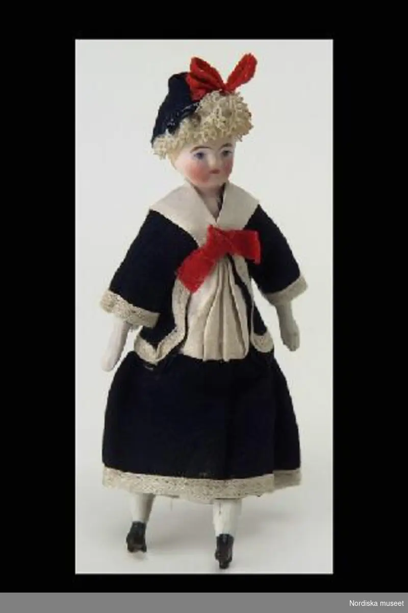 Inventering Sesam 1996-1999:
H 13,5 cm
Damdocka med huvud med sköld, halva armar och ben av biskvi, stoppad kropp, måþlade ansiktsdrag, målade svarta skor med klack, gjutet modellerat gulmålat hår, klädd i mörkblå matrosklänning av bomull med vit krage och garnering, hatt med spetsgarnering, röd rosett på hatten och i klänningens ringning.
Har tidigare hängt som dekoration i julgranen. Se även 210.044.
Helena C 1997