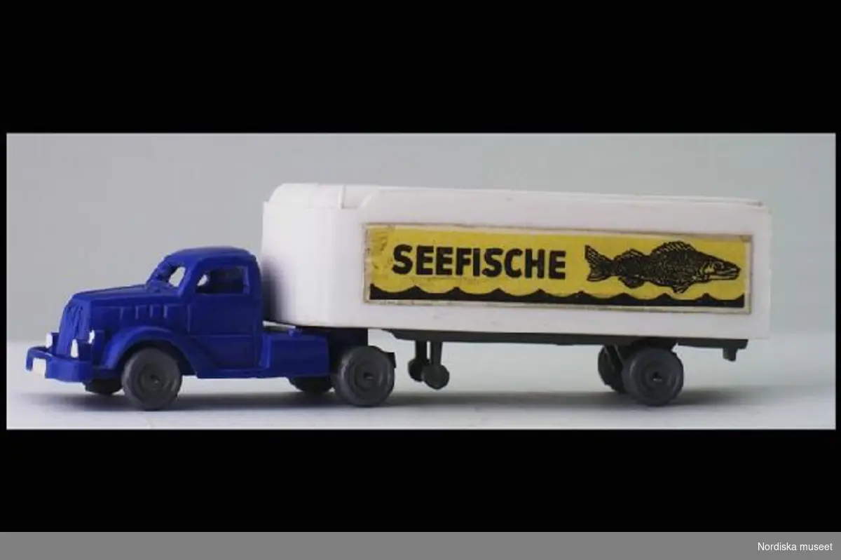 Inventering Sesam 1996-1999:
L 11,7   B 2,8  H 3,5 ( cm)
Dragbil samt släpvagn/trailer. Dragbil i blå plast, ihålig, öppna fönster, grå plasthjul. Släp i vit plast, markerade ej öppningsbara dörrar i bak. Påklistrade dekaler på sidorna med texten: "SEEFISCHE" och en bild av en fisk och böljande vatten. Saknar märkning. Enligt bilaga inköpt 16/4 1952 av givaren för 2,95 hos Eskader, Stockholm. Givaren, samlare av leksaksbilar 1947-1952, se inv nr 263.905 - 264.120.
Bilaga
Helena Carlsson 1996