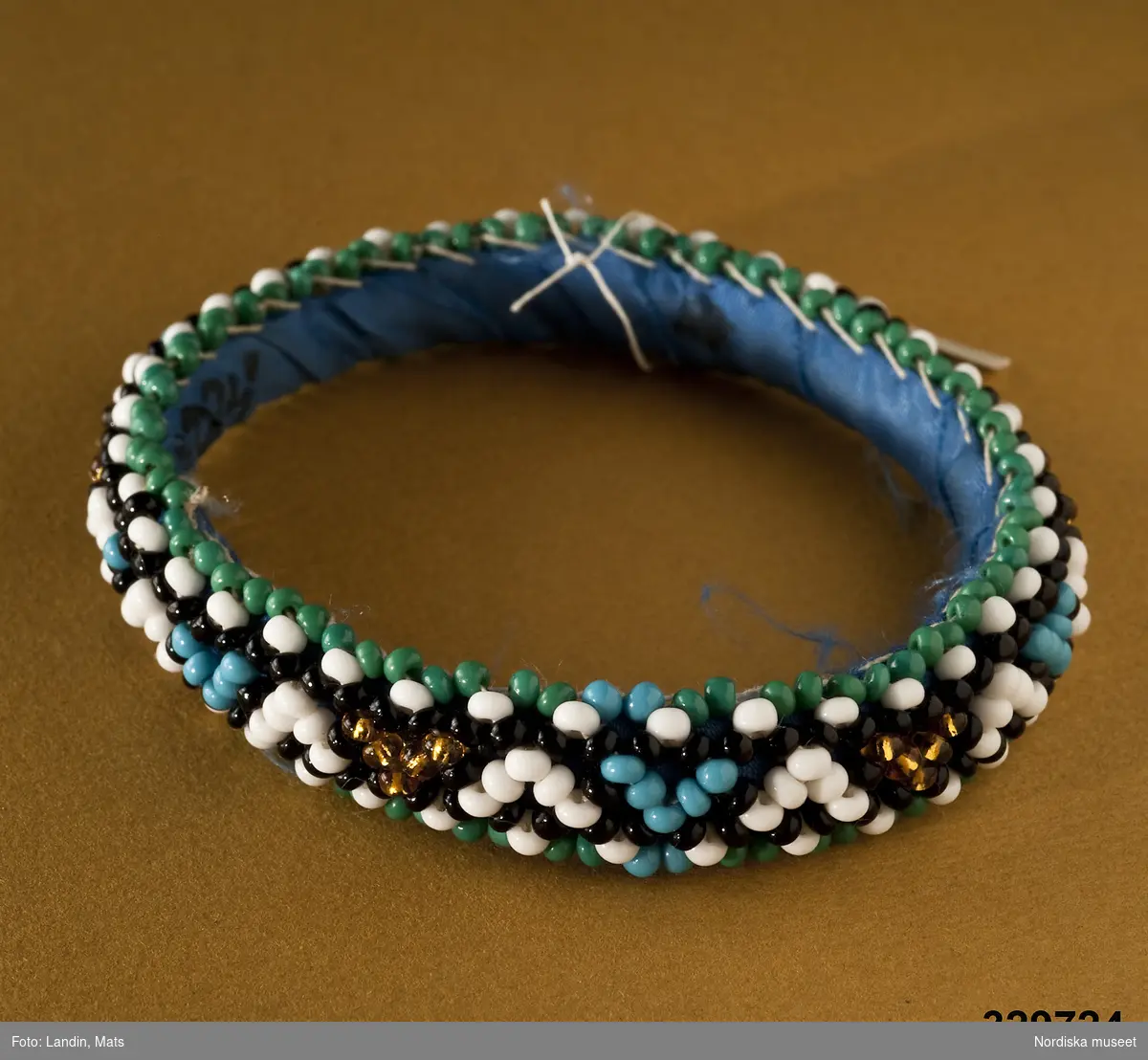 Runt armband prytt med små plastpärlor. Mönster i turkost, vitt och guld på svart botten. Fodrat med blått bomullstyg. Afrikanska influenser.
/Zingoalla Rosenqvist 2009-02-05