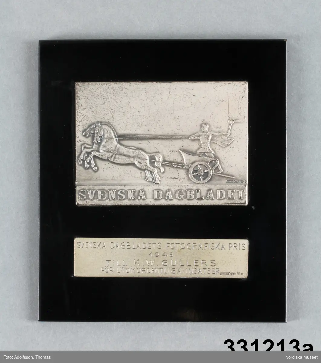 a) Medalj
b) Etui

a) Medalj/plakett, rektangulär svart platta av konstmaterial. På framsidan infattat två rektangulära silverplåtar. Den övre med relief i form av romersk krigare med fackla åkande i vagn dragen av två hästar och text "SVENSKA DAGBLADET". Den nedre silverplåten graverad text "SVENSKA DAGBLADETS FOTOGRAFISKA PRIS / 1948 / TILL K. W. GULLERS / FÖR UTOMORDENTLIGA INSATSER."  Bägge silverplåtarna med silverstämplar, den övre: "S&Co / s:t Erik / T8 / kattfot (tre kronor) / S." Den nedre likadana stämplar förutom årsstämpel, som är  "Y8" istället. På baksidan liten triangulär hängare i metall, runda hål för kramporna som håller fast silverplåtarna
/Leif Wallin 2011-01-21