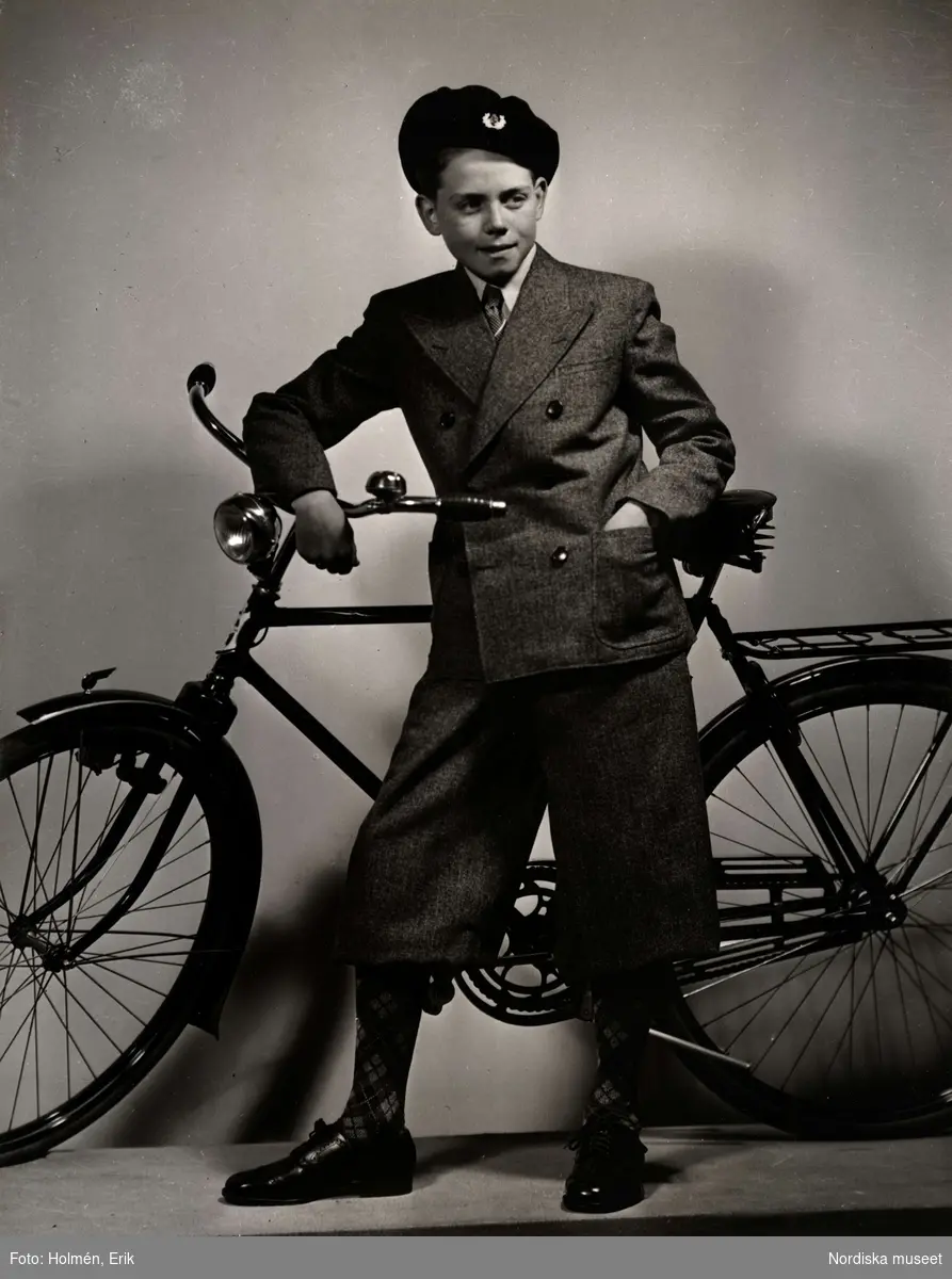 Barnkläder visas på Nordiska Kompaniet 1941. Pojke poserar vid cykel, iklädd kavaj och knäbyxor av tweed, rutiga strumpor, promenadskor och basker.