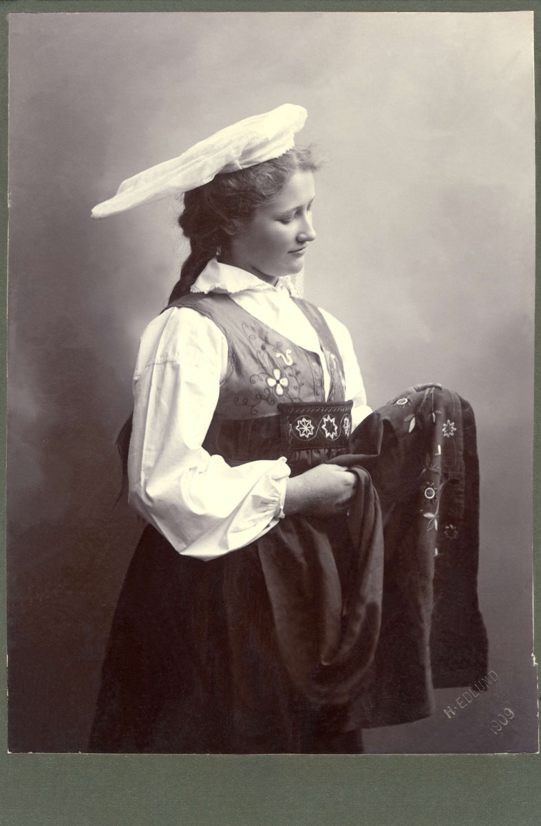 En kvinna står och studerar sitt förkläde till folkdräkten. Klädd i högtidsdräkt från Österåker som den bars vid sekelskiftet 1900.