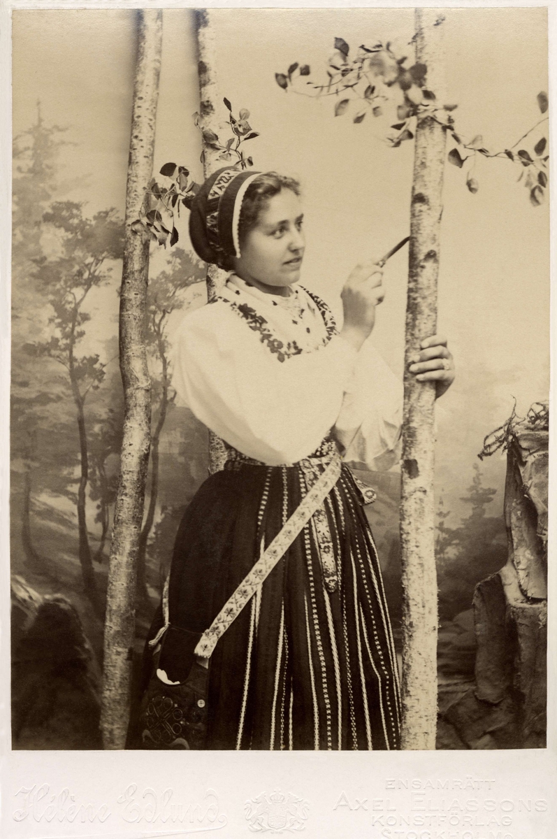 En kvinna ristar i ett träd klädd i Leksandsdräkt som den bars kring 1900.