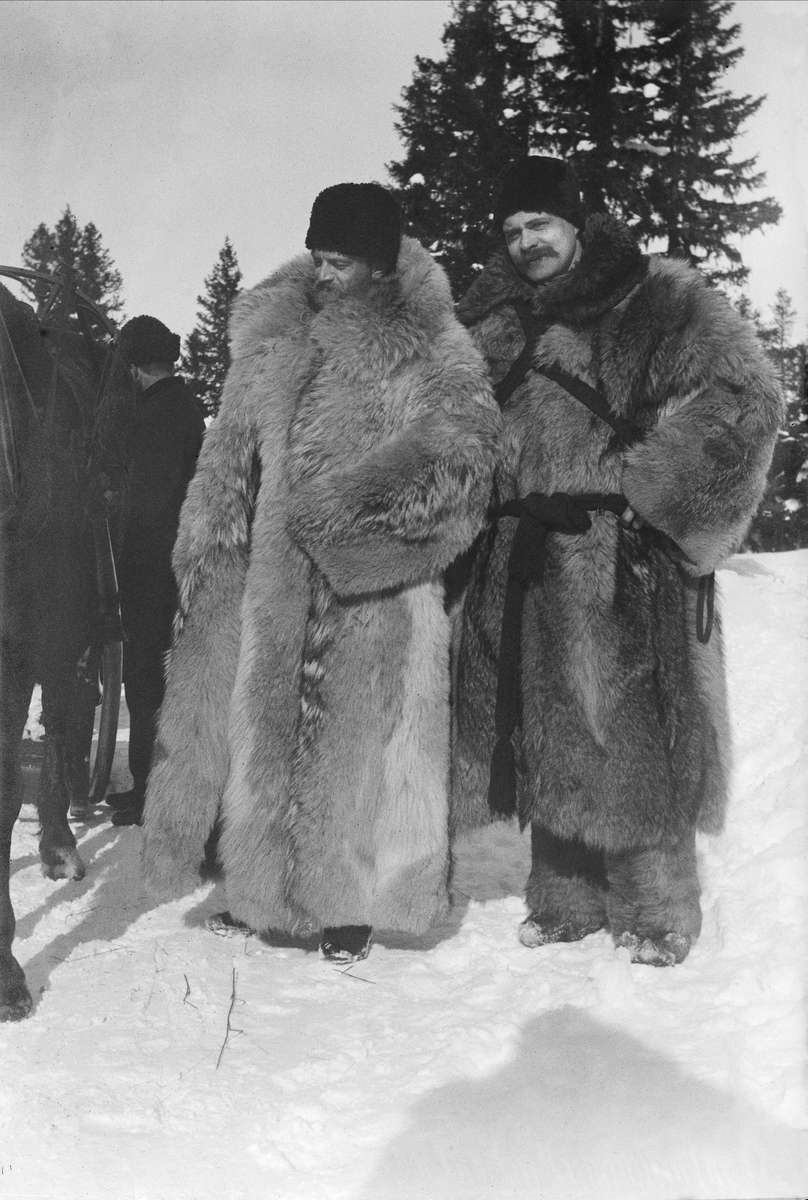 Två män i vadlånga pälsar står i snöigt landskap, Umeå.