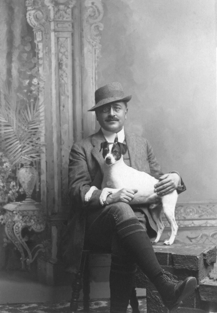 Ateljéporträtt av löjtnant Arthur Bäckström sittande iklädd knäbyxor, kavaj och hatt med hunden Dono i famnen.