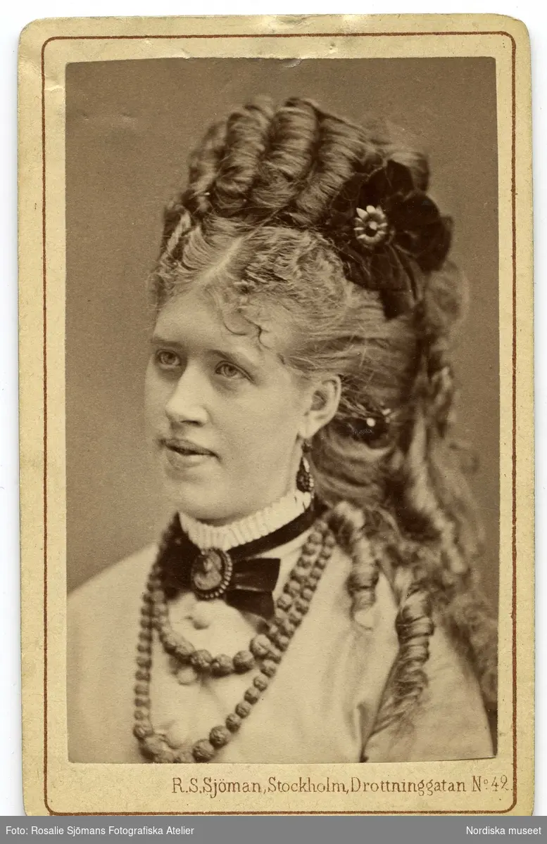 Porträtt av ung kvinna med hög, konstfärdig frisyr med en sammetsblomma som hårsmycke. Kvinnan har en vit skjorta med veckad ståkrage, sammetsband med en kamé runt halsen samt ett pärlhasband och hängande örhängen. 1870-tal.