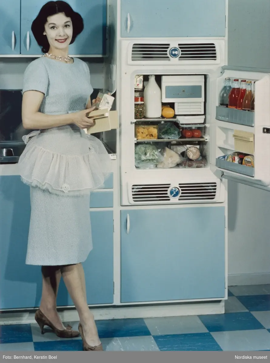 Kök i blått. Kvinna vid öppet kylskåp med diverse matvaror. 1950-tal.
