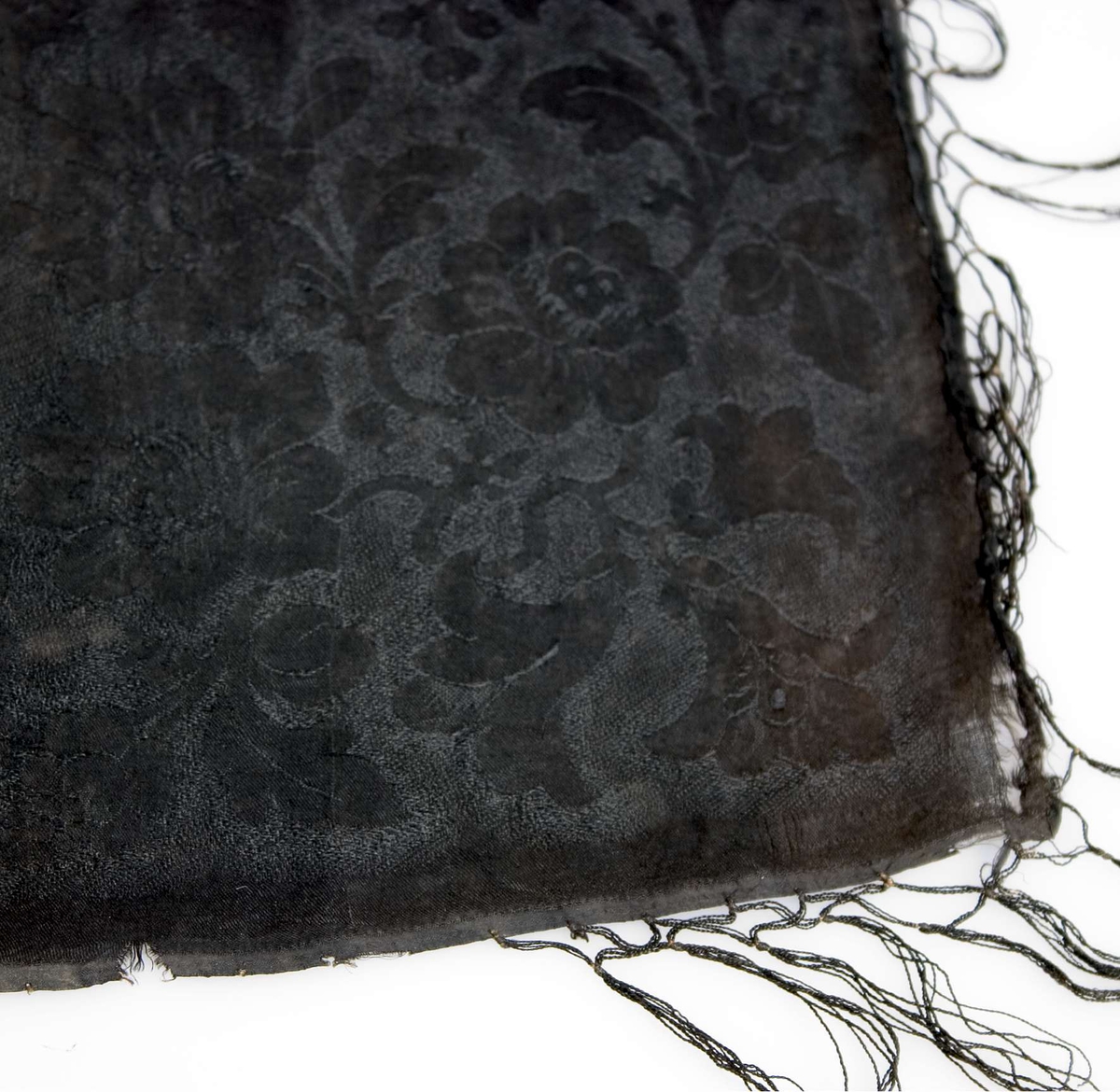 Huvudkläde av svart mönstervävt siden. Blommönster över hela ytan. Svarta iknutna silkefransar. Fållad för hand i två sidor.