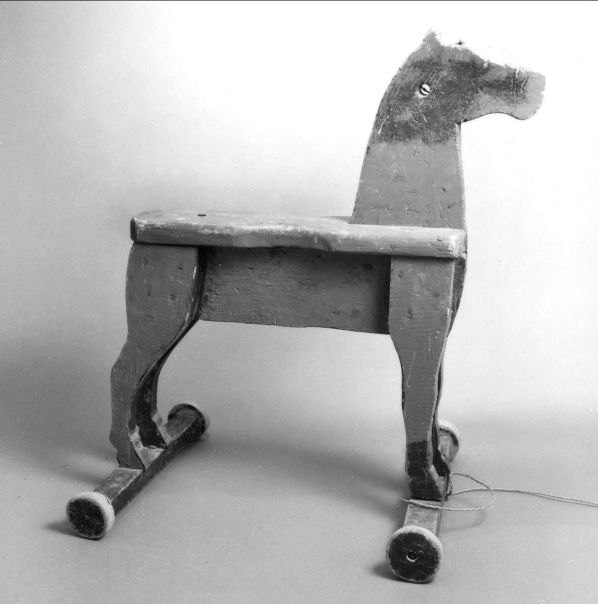 Häst av trä, tillverkad av brädor, står på två tvärställda brädor med hjul av trä i båda ändar. 