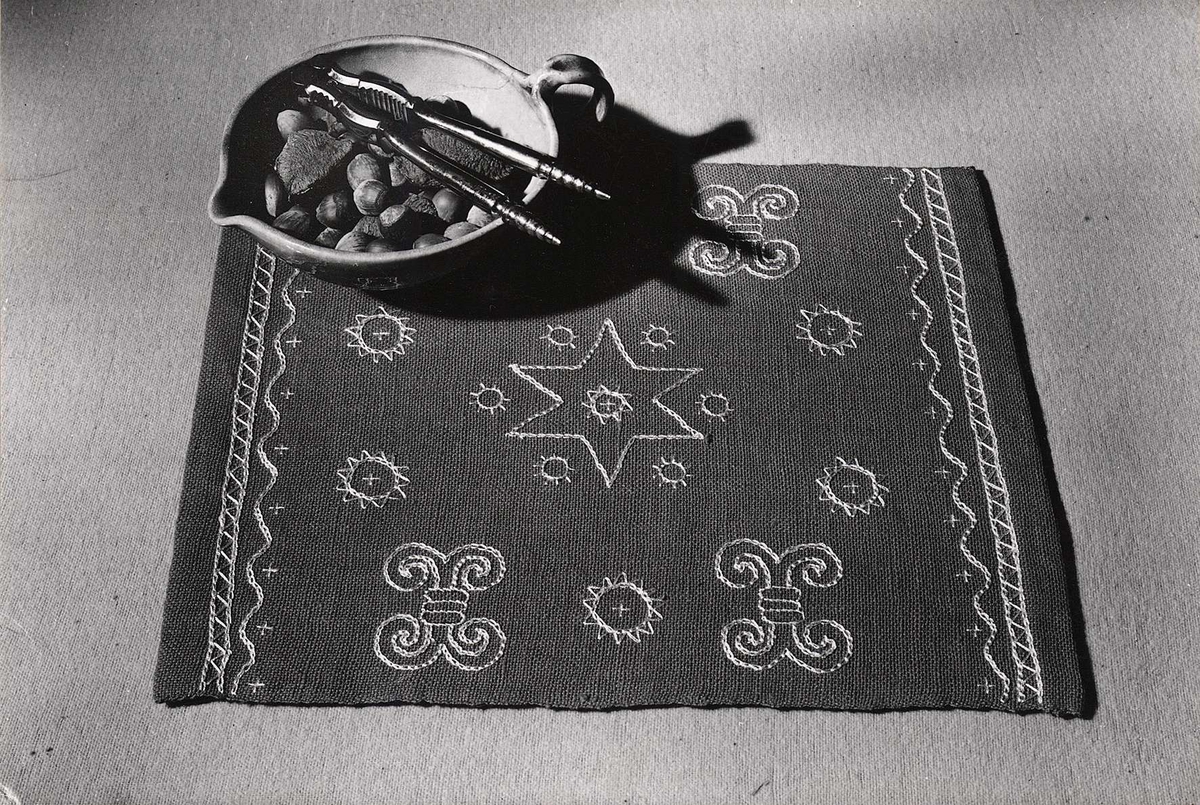 Två svartvita fotografier klistrade på ett 22 x 27 cm stort brunt kartongblad. Det ena fotografiet visar hela duken och det andra visar en detalj av dukens broderi.
Kartongbladet är märkt B-2162.