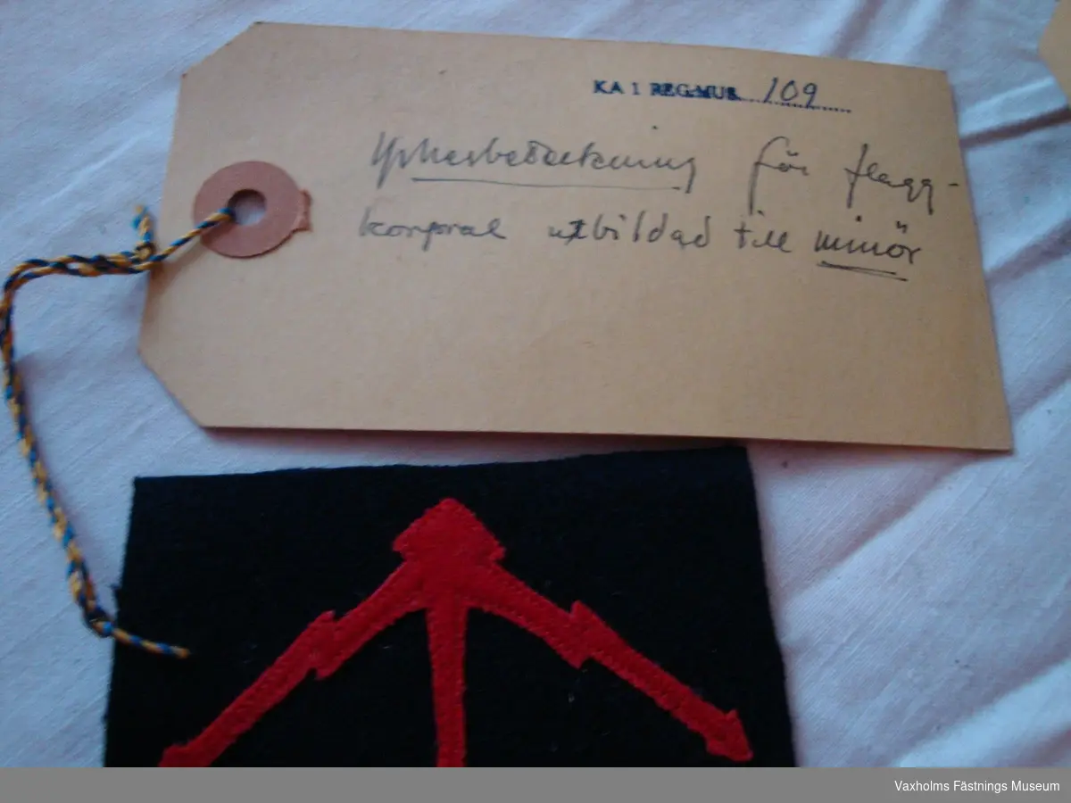 Yrkesbeteckning för flaggkorpral utbildad till minör. Tre viggar i ett knippe, av rött kläde i applikationssöm på mörkblått kläde.