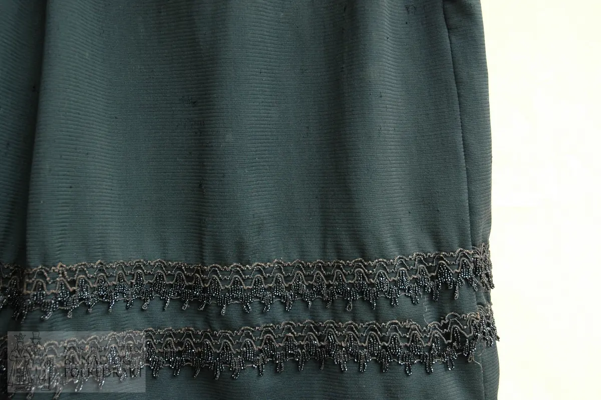 Del av to- delt kjole i svart, forhaldsvis tynt ulltøy. Skjørtet er sydd saman av tre høgder, samansydd på maskin. Brei fold framme med splitt i venstre side(saumen). 5 folder som går bakover på kvar side til dei møter eit rynkeparti mb. Rynkene har eit ekstra tøy på innsida av blomstra bomull, to parallelle rynketrådar. Smal linning av kypertvove bomull som blir hekta saman ved splitten på venstre side. Nederst på skjørtet er det påsydd to lengder av blonde med perler på. Skjørtet er skonert nederst med beige bomullslerret.
