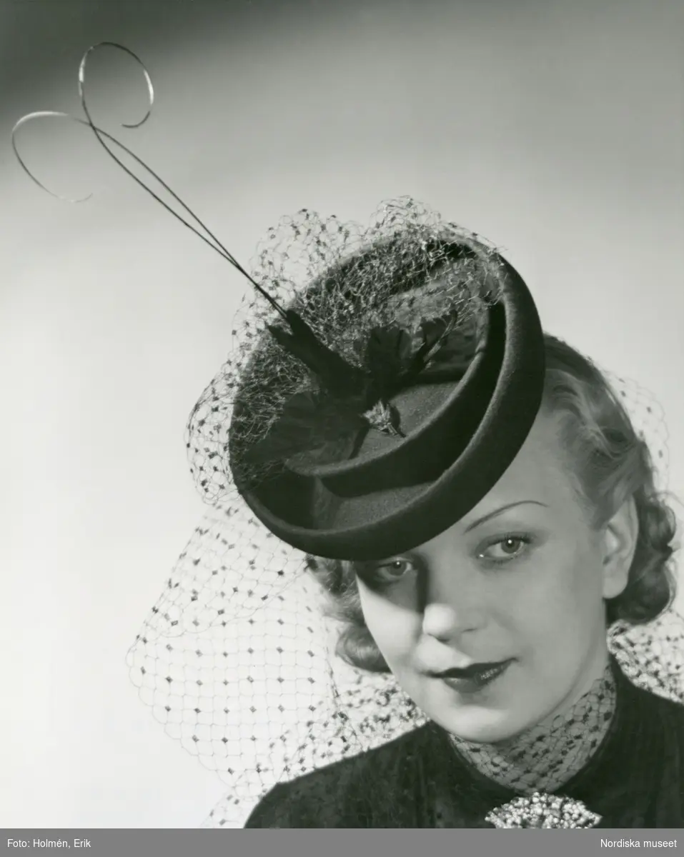 Porträtt av kvinna i hatt med flor och fjädrar, mörk blus och brosch.