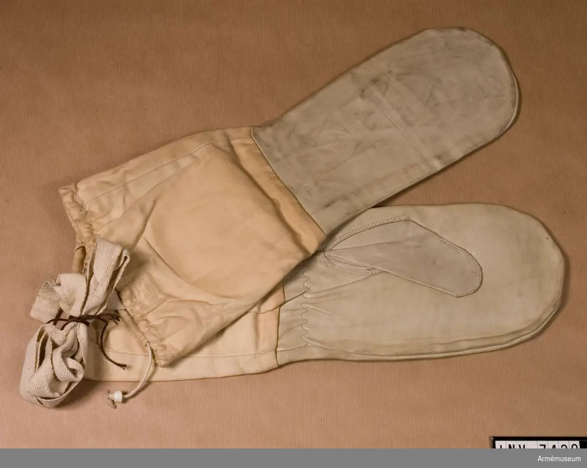 Skidhandskar arbm/1952. Vita läderhandskar med långa tygskaft och långa vita band för att de ej skall tappas eller blåsa iväg vid skidåkning. Skaften är ihopdragbara vid överkanten med resårband.