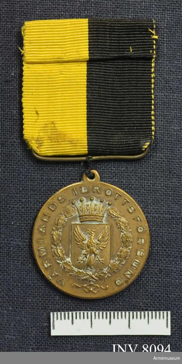 Samhörande AM 8076-.
Diameter 30 mm. Tjocklek 2 mm. Vikt 25 g. Färg brons (kopparbrun). I relief på åtsidan (adversen) i mitten Värmlands vapen och i kanten står det: Värmlands idrottsförening. Frånsidan (reversen) är ograverad, har endast lagerkransen i kanten. Medaljen hänger i ett svart och gult ripsband 35 x 35 mm. 
