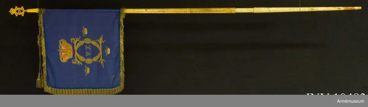 Grupp B I:2.

Duk av mörkblå (indigoblå) 4-skaftat fansiden med broderade emblem: CXVC. Rikt profilerade C under kunglig sluten krona med rött inner- och ytterfoder och silverstenar. Tre kronor med blått innerfoder i två nyanser. Alla broderier i guld. Kronans  foder rött fansiden med schattersöm. Frans av grövre och finare guldkantiljer. Signerad 18 3/5 64. Fastspikad med  guldbeläggningssnöre (majorssnöre). Kupiga tännlikor.
Stång: Rött med guld ovan duken, ränna för kravatt, doppsko  saknas. 660 mm, 1620 mm, 1780 mm, 2670 mm. Spets med Karl XV:s krönta monogram, l 152 mm. Holk 80 mm. Ränna 120 mm. Kravatt av gespinsterarbete, tofsar som fransen. Enkla knutar. Längd 1270 mm.