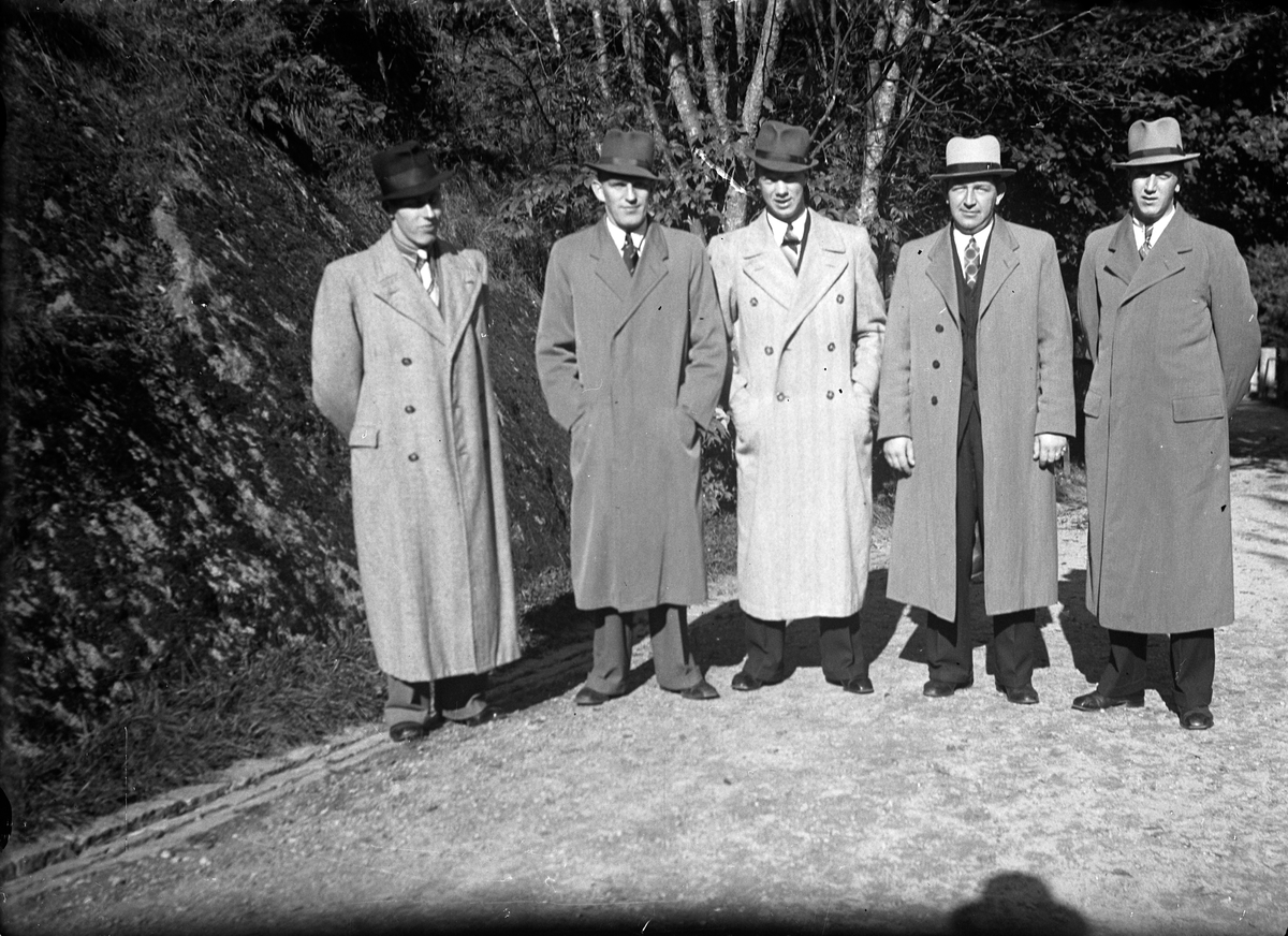 Fem män klädda i rock och hatt står på en väg, vid sidan syns en bergssluttning och träd.
