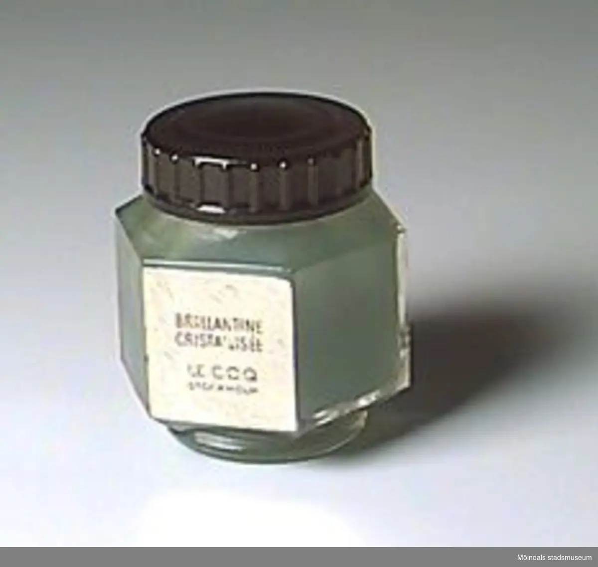 Sexkantig glasburk med rund fot, svart skruvlock. Innehåller grön pomada. Etikett i guld: "Brillantine Cristallisée/ Le coq/ Stockholm". Pris enligt prislapp: 4,75 kr.Kjellberg arbetade i väveriet på Mölnlycke fabriker 1916-1969.