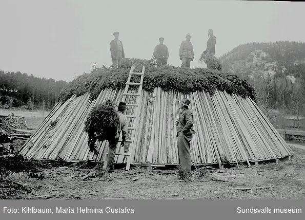 Män bygger en kolmila. Troligen vid ett sågverk eftersom materialet består av bräder.