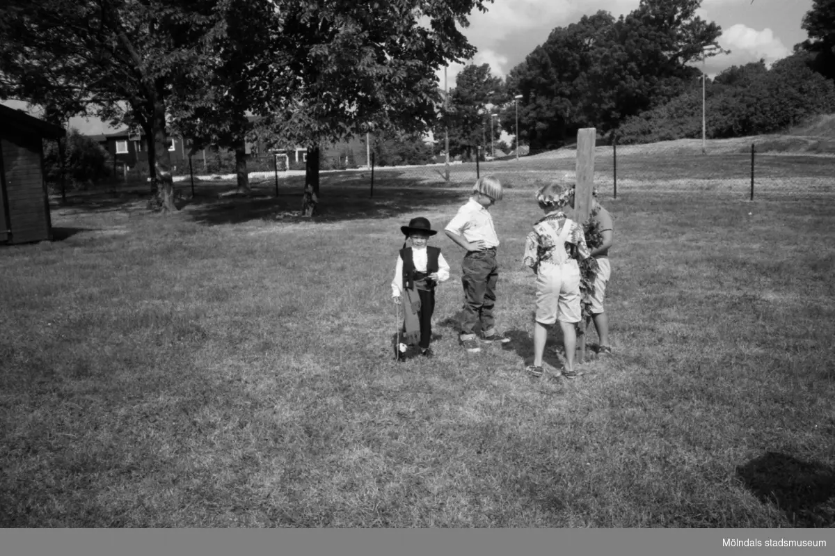 Fyra barn från Katrinebergs daghem står utomhus på en stor  inhägnad gräsmatta. Ett litet barn, iklädd hatt, väst och käpp, går mot fotografen. Ett par av barnen håller en stående träplanka. I bakgrunden ser man några hus samt höga träd.