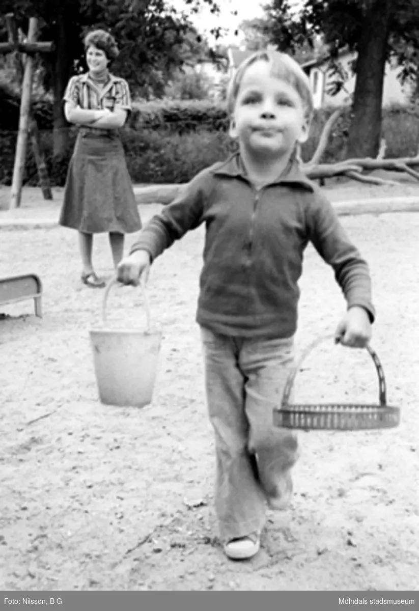 En pojke leker i sandlådan och fröken ser på i bakgrunden. Holtermanska daghemmet 1973.