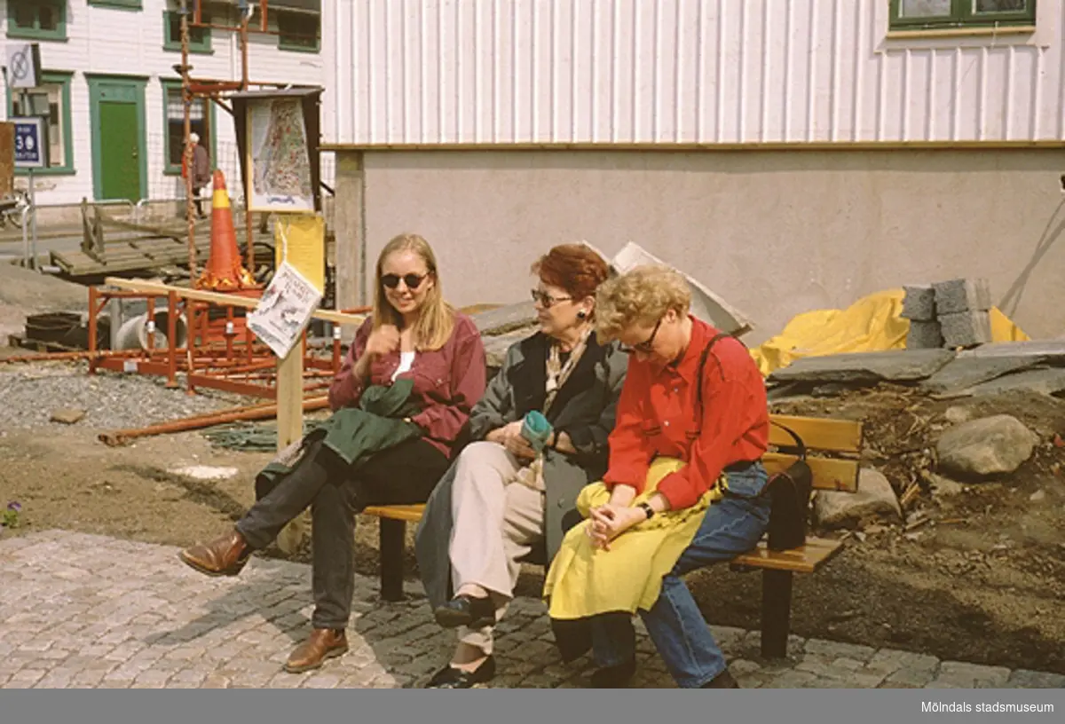 Tre kvinnor sitter på en parksoffa vid Lyktplatsen, Kvarnbydagen 25/4 1993. Gatlyktan (ej i bild) invigs. Kråkans Krog (vitt hus med grön dörr) ses till vänster.