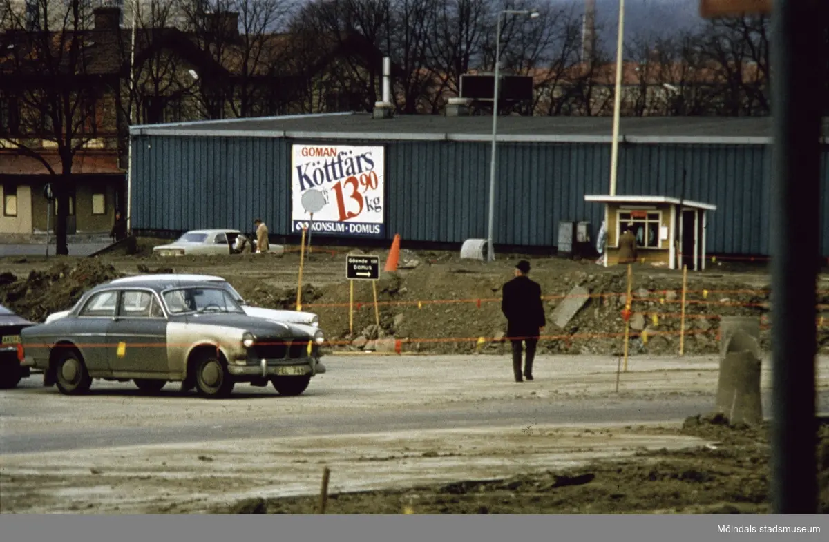Domus barack vid Nya Torget i Mölndal, 1970-tal. Främst ses parkerade bilar uppställda på en parkeringsplats. Till höger ses en liten kiosk.