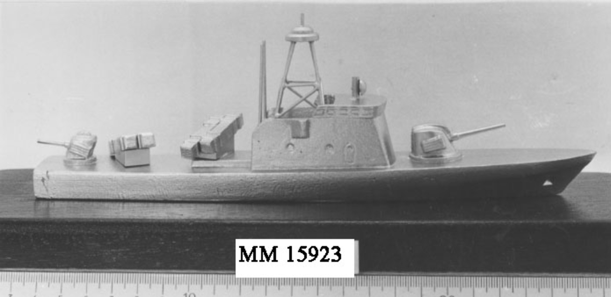 Modell av torpedbåt, typ Spica, Båten är silverfärgad. På fördäck sitter en kanon liksom en på akterdäck. Bakom överbyggnaden är två robotramper placerade, en åt styrbord och en åt babord. Hela modellen är påklistrad på en bottenplatta av mörkbrun mahogny.