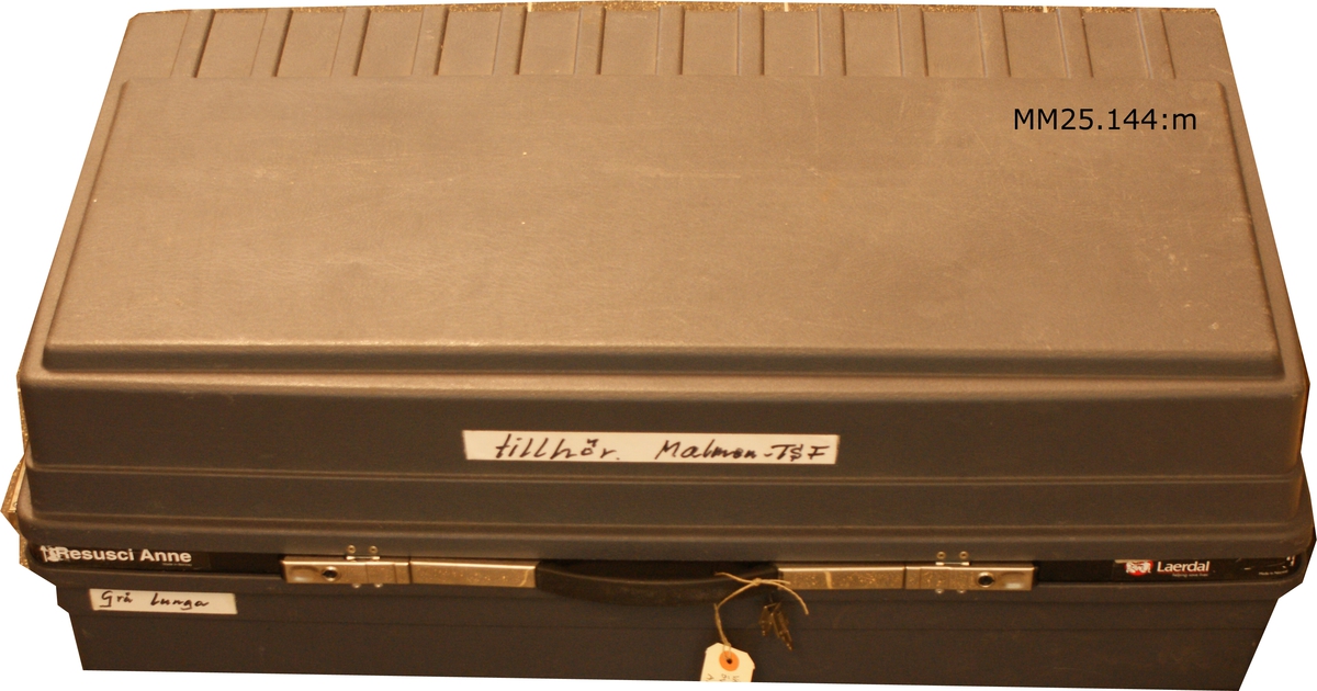 Grå resväska med hjul. Märkt med svart tusch på vit tejpremsa: "Tillhör Malmen TSF". (TSF betyder sannolikt Truppserviceförråd).