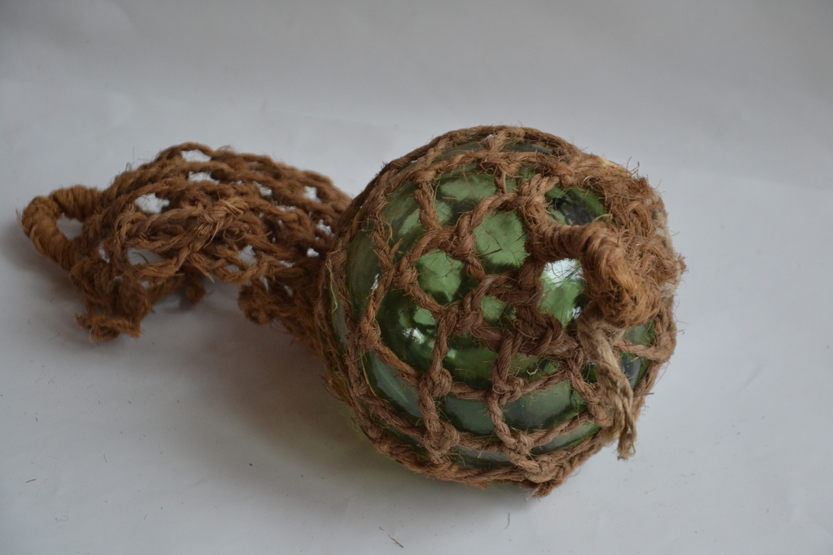 En grønn glasskule med nett av kokostau festet rundt seg. En hempe av kokostau er festet i den ene enden. På glasskula er merket bokstavn M. Nettet er tvinnet på plass, ikke knytt.