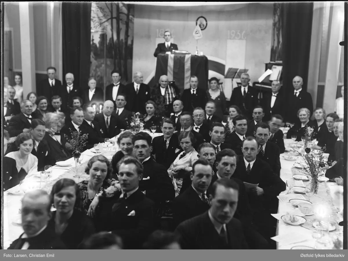 Gruppe menn og kvinner med festpyntet langbord, scene med taler og vimpel, tekst bak: 1836-1936.
Ukjent forening
