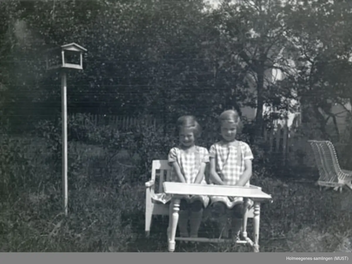 To små jenter på en liten benk i hagen på Holmeegenes i Stavanger. Et fuglebrett vises til venstre i bildet.