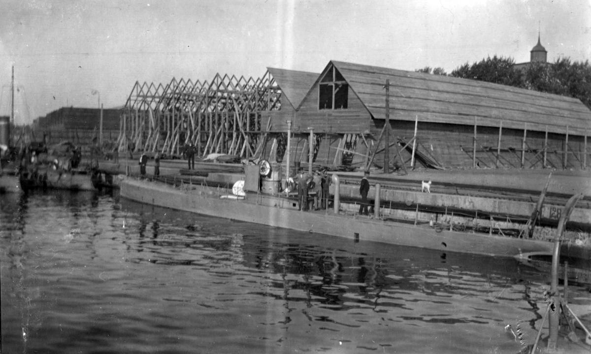 Karlskrona 1914. Foto på undervattensbåten Nr: 4 förtöjd vid brygga. På ubåten och kajen står några sjömän. I bakgrunden syns byggnader under uppbyggnad.