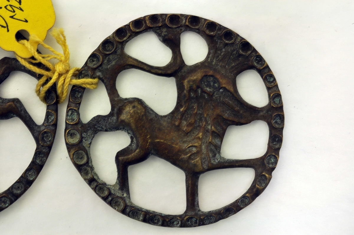 Fra protokoll: 1 par beslag av bronse (legeringsmetal). Cirkellaga sylgjebeslag i gjenombroti støypearbeid. Motiv: heraldisk løve innskrivi i cirkelring. Tvermaal 4,25 cm.