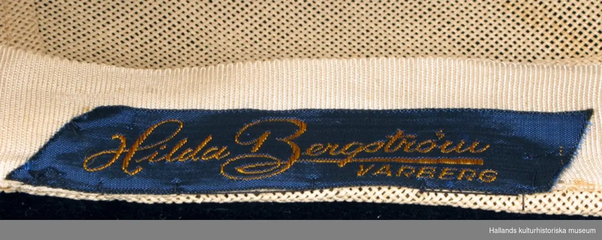 Damhatt/basker av benvit strå. Barettmodell, hög i vänster sida, rund platta som sluttar åt höger. Odekorerad.
Märkning inuti hatten, "Hilda Bergström VARBERG", namnet i guld på blå bakgrund.