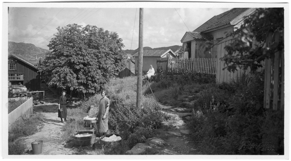 'Nedgång till ''Söderns'' tilläggsplats. Till vänster Hermanssons gård, t.h Albertina Hellmans. 2 kvinnor i förgrunden, den ena vid tvättkar och den andra med blombukett i handen. På farstutrappan till huset till höger står 1 kvinna och 1 person sitter på trappa nedanför huset. Kläder. Klädsel. ::  :: Serie fotonr 1888-1963.'