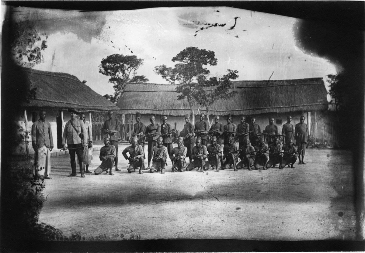 'Diverse fotografier från bl.a. dåvarande Nordrhodesia, nu Zambia, tagna av Konsul Magnus Leijer. ::  :: 2 rader av män. Främsre raden består av 11 st män som sitter på huk. Bakre raden består av 14 stående män, alla med vapen. Bredvid står 2 män.'
