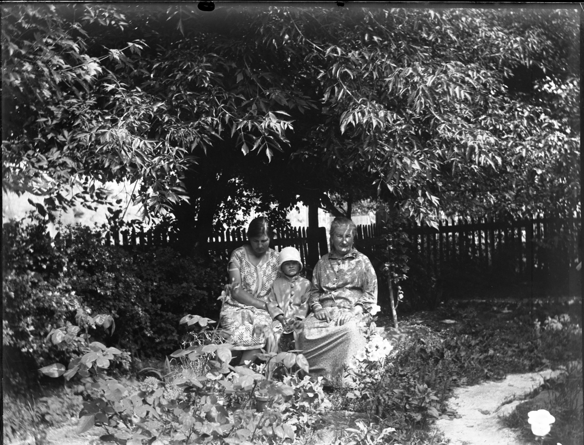 'Bildtext: ''Kyrkesund 1929.'' :: 2 kvinnor och 1 barn sittande på träbänk i en trädgård, trästaket i bakgrunden. ::  :: Ingår i serie med fotonr. 5233:1-14'