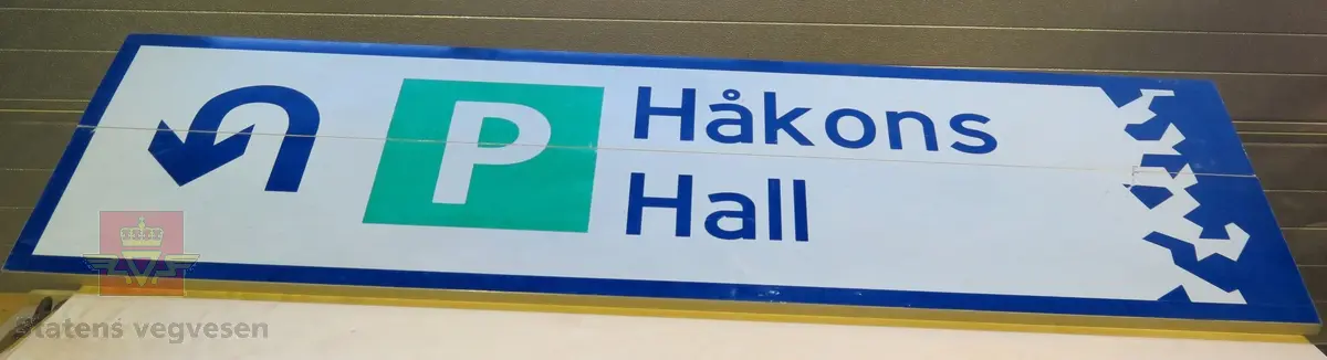 Rektangulært skilt av aluminium. Har symbol for parkering og kjøreretning samt teksten "Håkons hall". Flerfarget, med grå bakside.