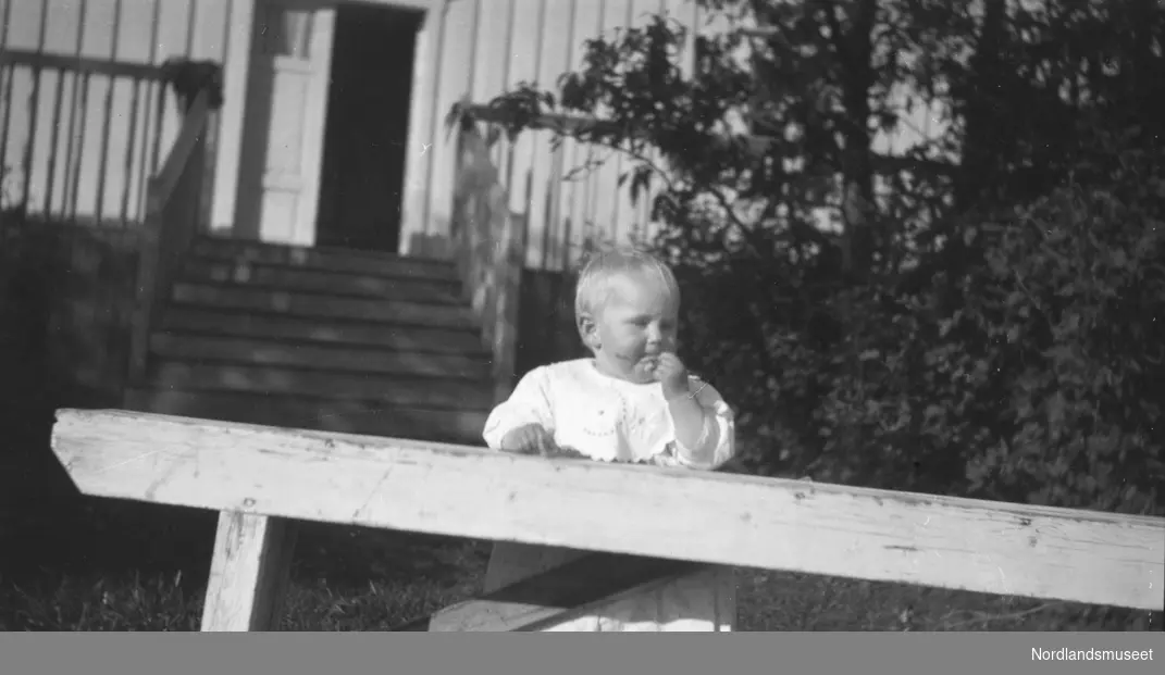 Bildet viser Unni Strand, 1917 som liten unge, står ved en benk. Et bolighus står i bakgrunnen.