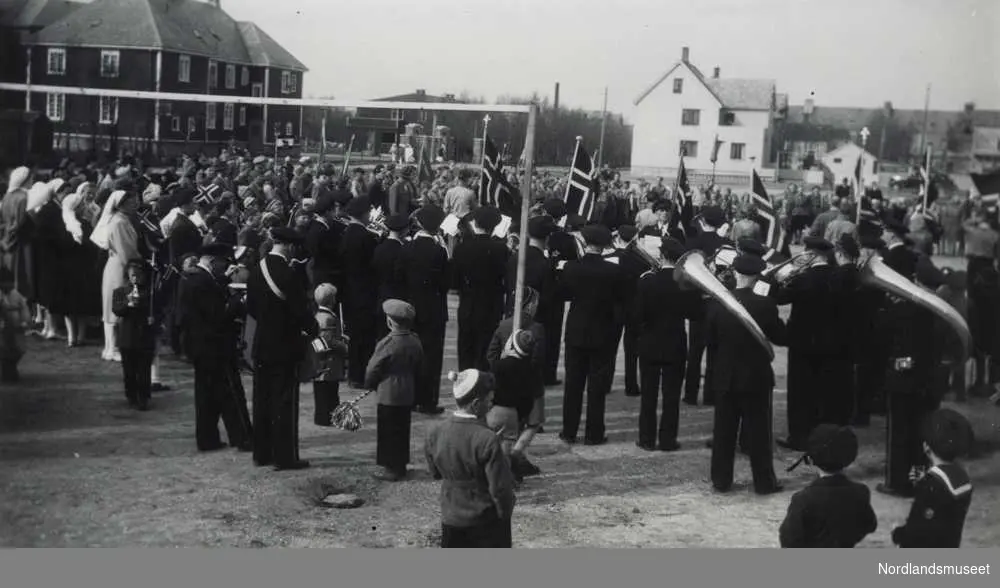 17. mai 1949 i Bodø. Folkemengde samlet på den gamle idrettsplassen. Hornmusikk og norske flagg. Bebyggelse i bakgrunnen.