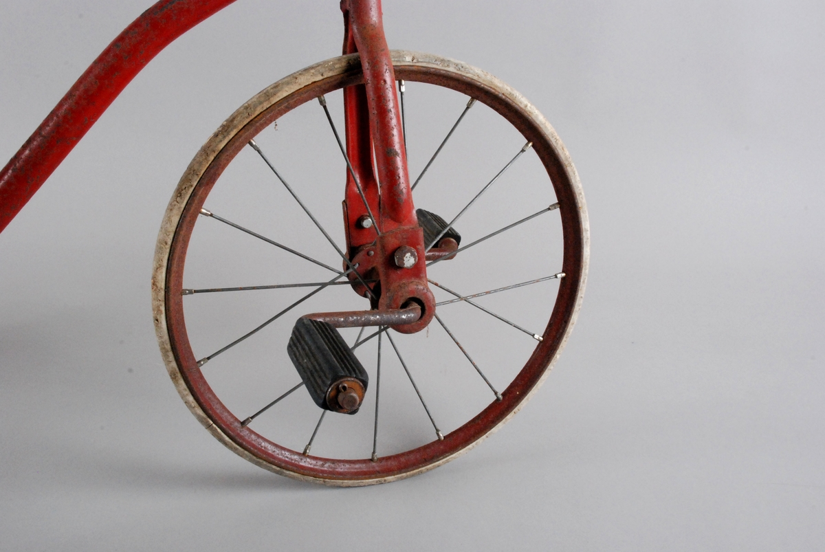 Trehjulsykkel i metall med gummihjul, gummi-pedaler og handtak i gummi. Sete trekt med sort bomullslerret.