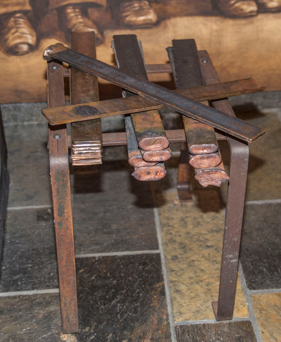 Flate jernplater montert sammen med stålnagler slik at de danner ett stativ med flat topp og fire bein. 15 teiner er montert på stativet.