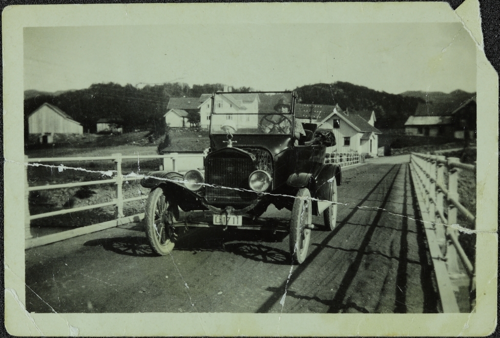 Bil (Ford T) kjørende over en bro, bak rattet en ung dame, ved siden av et barn