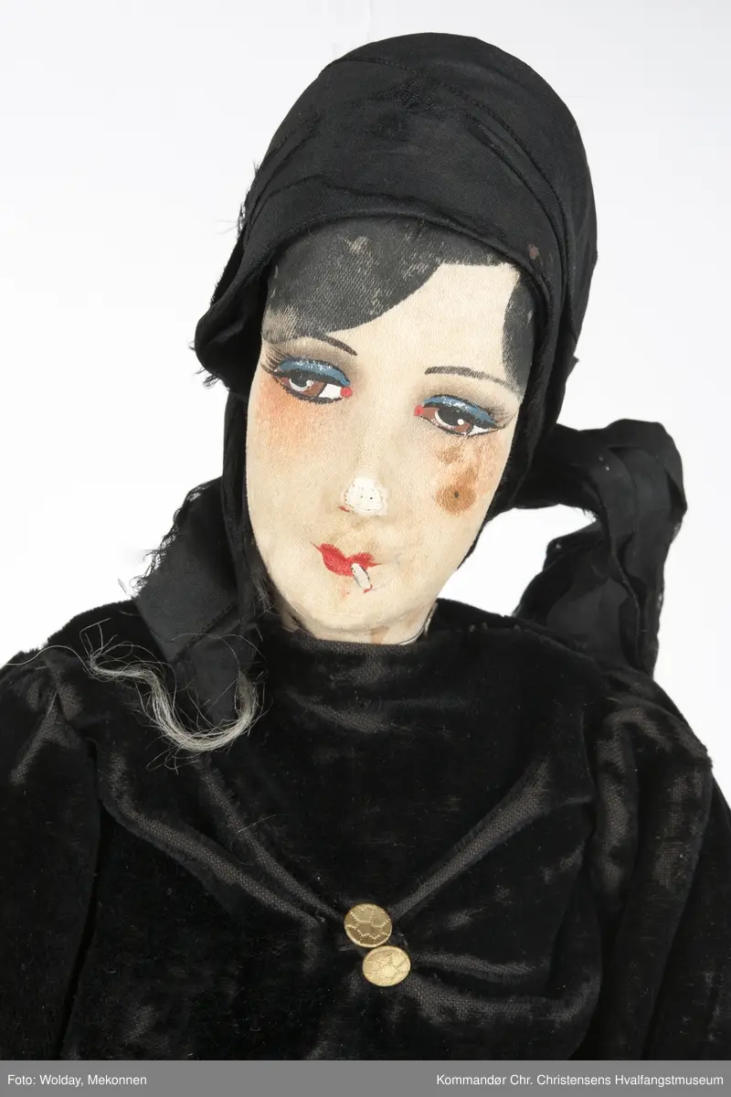 Dukken: sminket kvinneskikkelse med sigarett i munnen,  tørkle på hodet og sort todelt dress (Langermet, kort bluse, lange bukser, strømper.