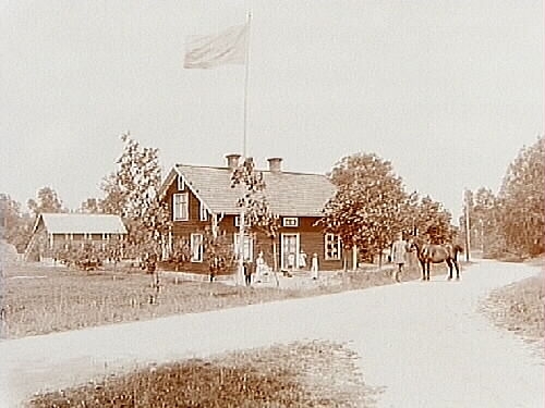 En och en halvplans bostadshus, handelsbod, 5 personer utanför byggnaden, en person med en häst på vägen.
Handlare A. Göransson