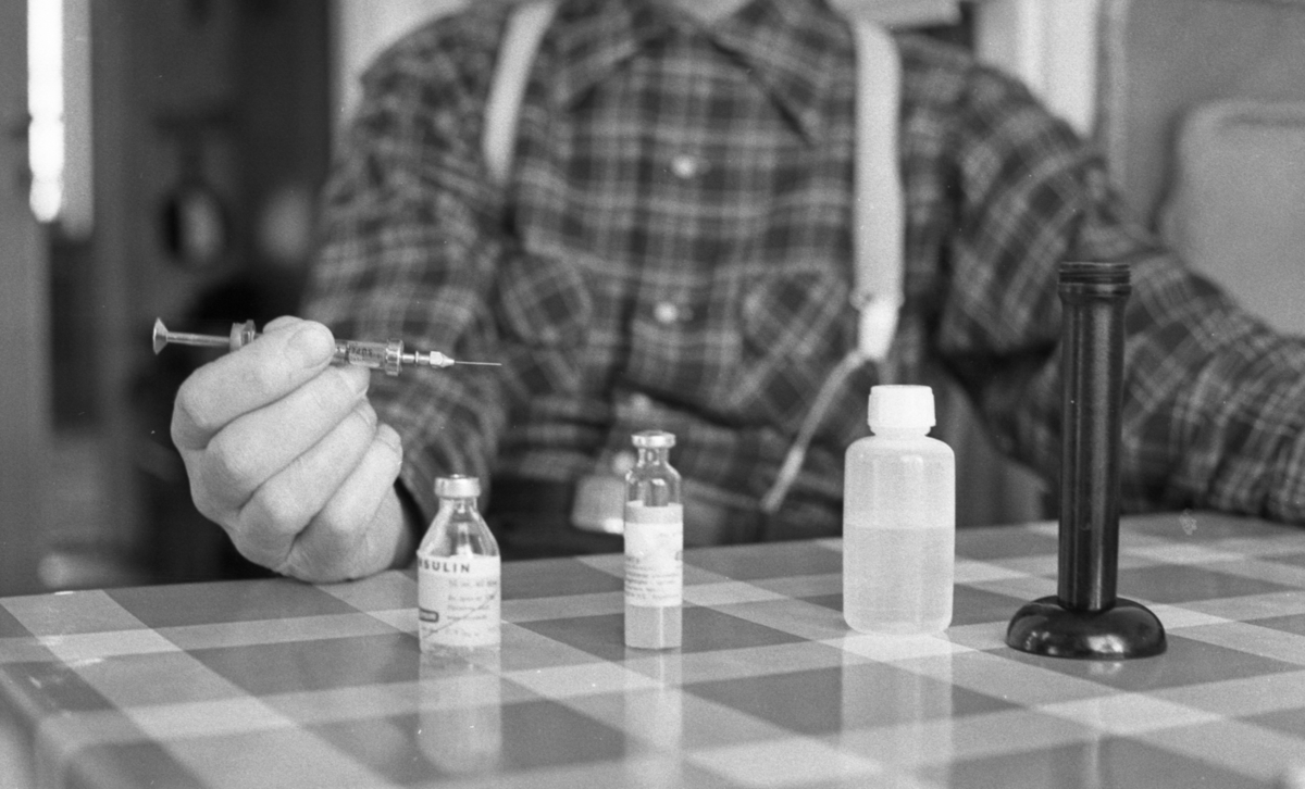 Diabetiker, 5 februari 1966

En man klädd i rutig skjorta, hängslen och byxor sitter vid ett bord och håller en spruta i sin högra hand. På bordet står en flaska med insulin samt två ytterligare flaskor. Även en avlång cylinderformad svart sak står på bordet.